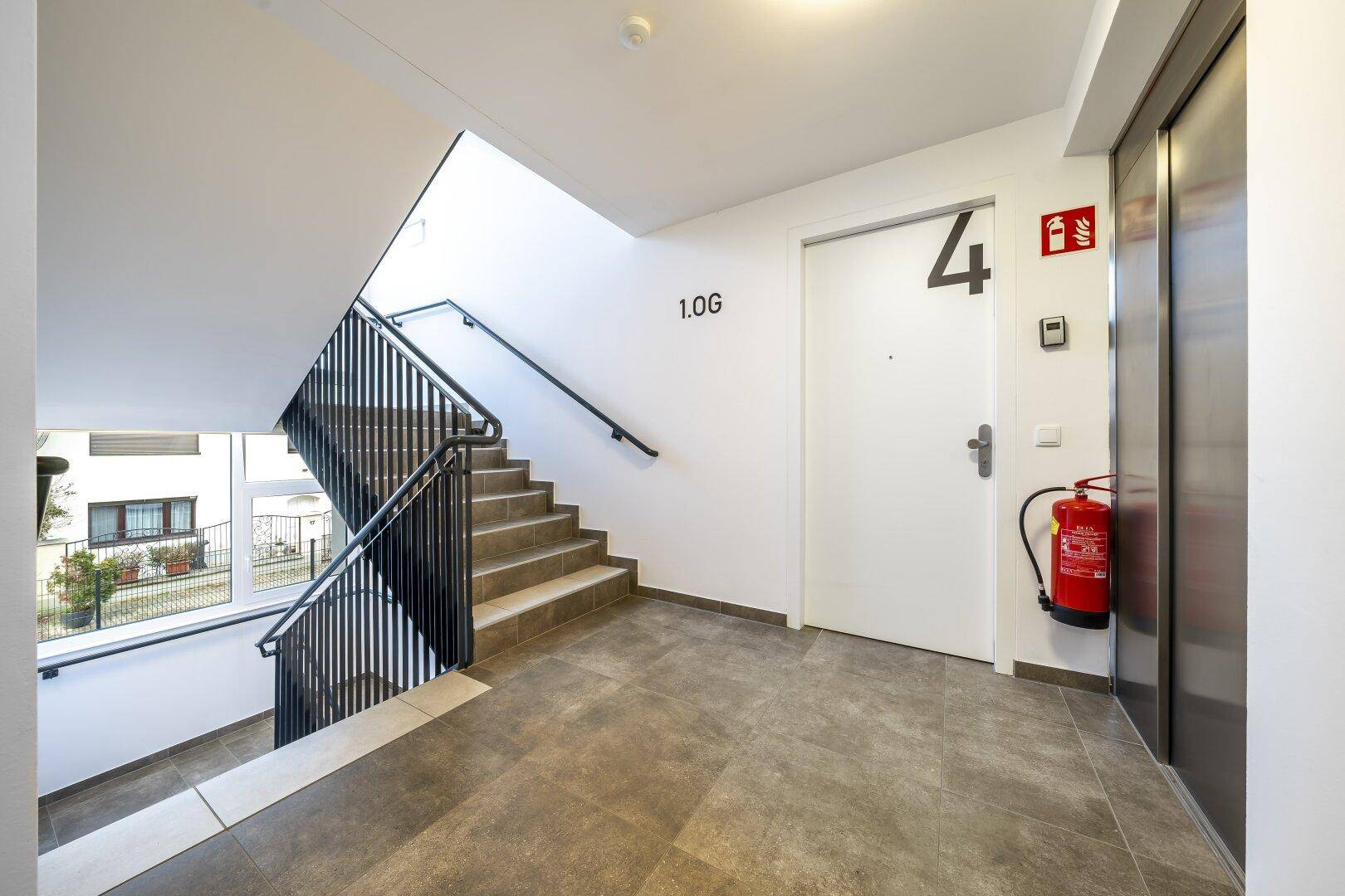 Treppenhaus mit Fliesenboden, Geländer, Aufzug und Zugang zu den Wohneinheiten.