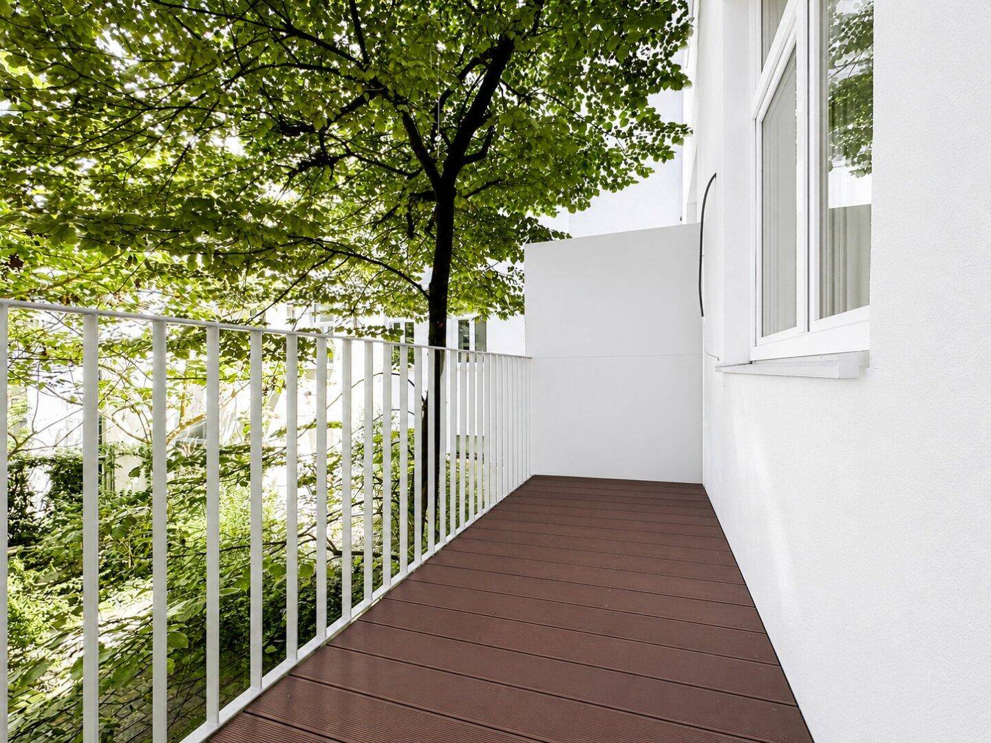Schmaler Balkon mit Holzboden und weißem Geländer, umgeben von grünen Bäumen.
