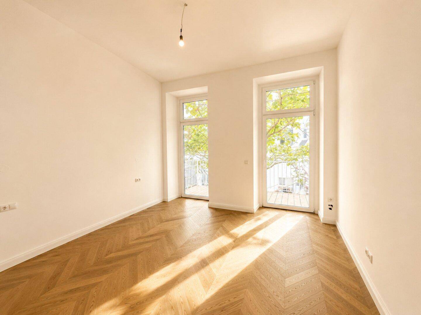 Sonniger Raum mit Fischgrätparkett und großen Fenstern, die viel Tageslicht hereinlassen.