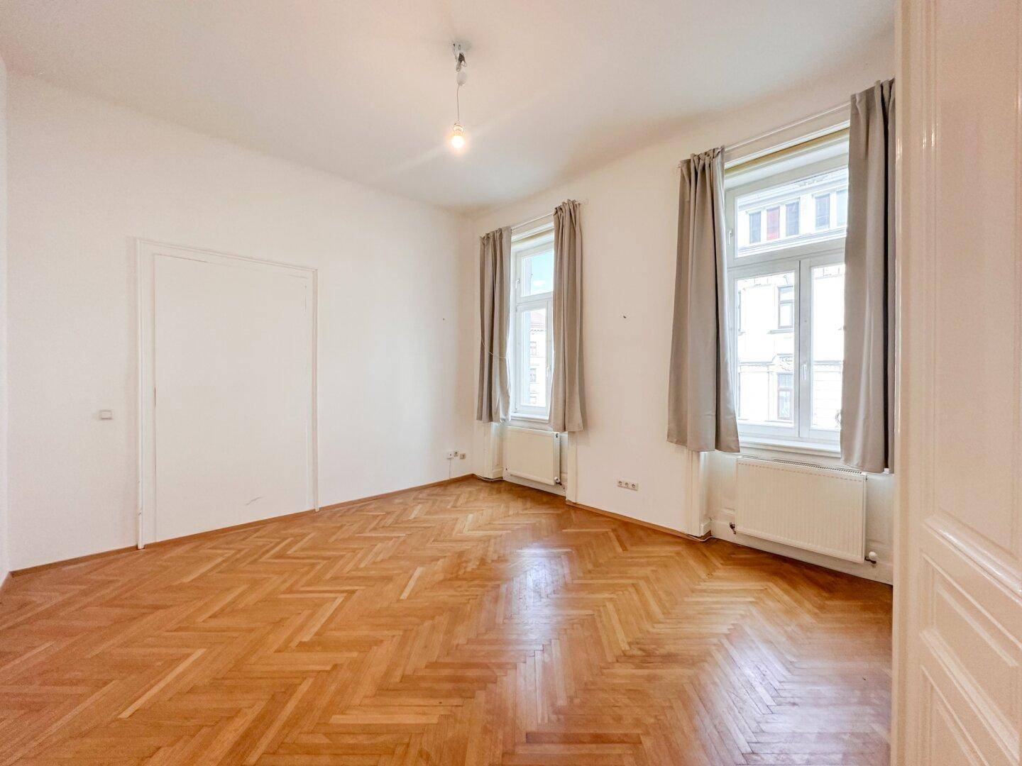 Lichtdurchflutetes Zimmer mit elegantem Fischgrätparkett und zwei Fenstern mit grauen Vorhängen.