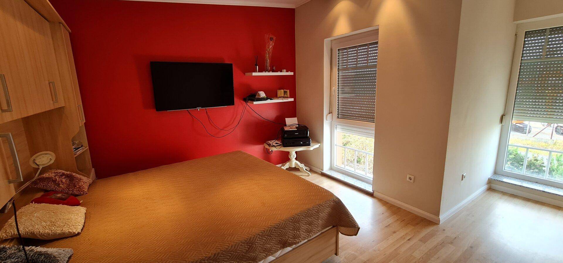 Modernes Schlafzimmer mit roter Akzentwand, Holzboden und Flachbildfernseher.