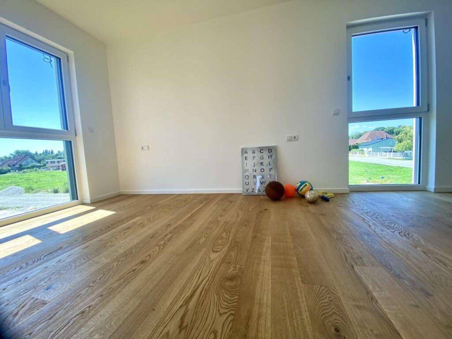 Helles, leeres Zimmer mit Eichenparkettboden und großen Fenstern mit Blick ins Grüne.