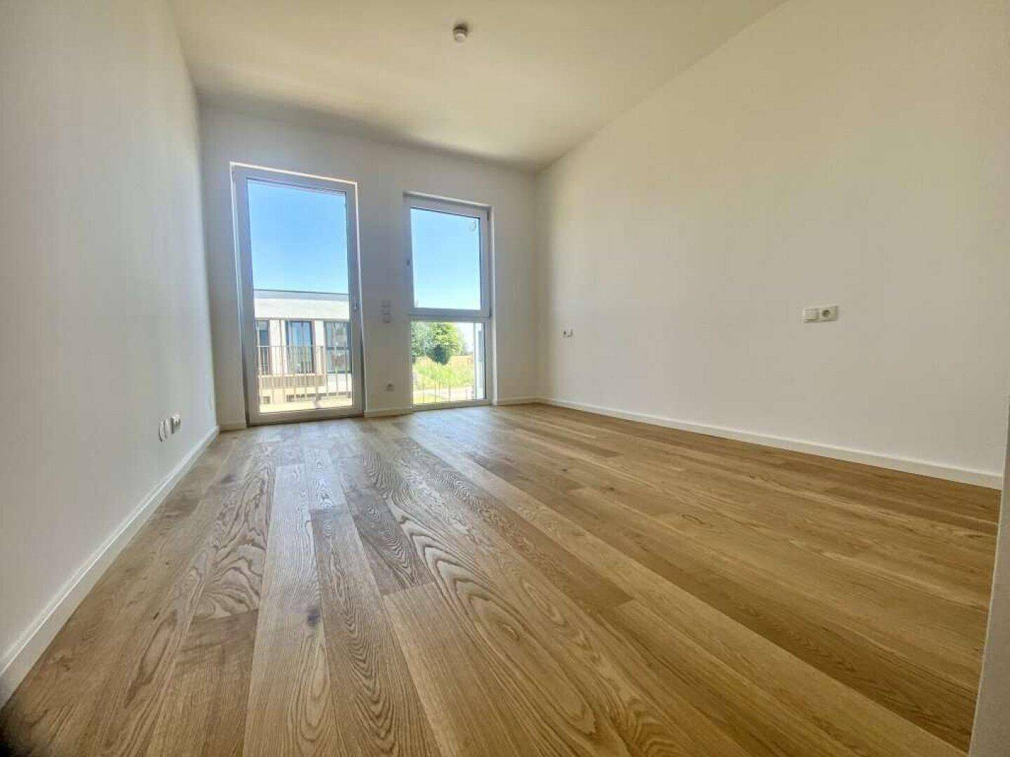 Leeres, helles Zimmer mit Eichenparkettboden und großen Fenstern mit Blick ins Grüne.