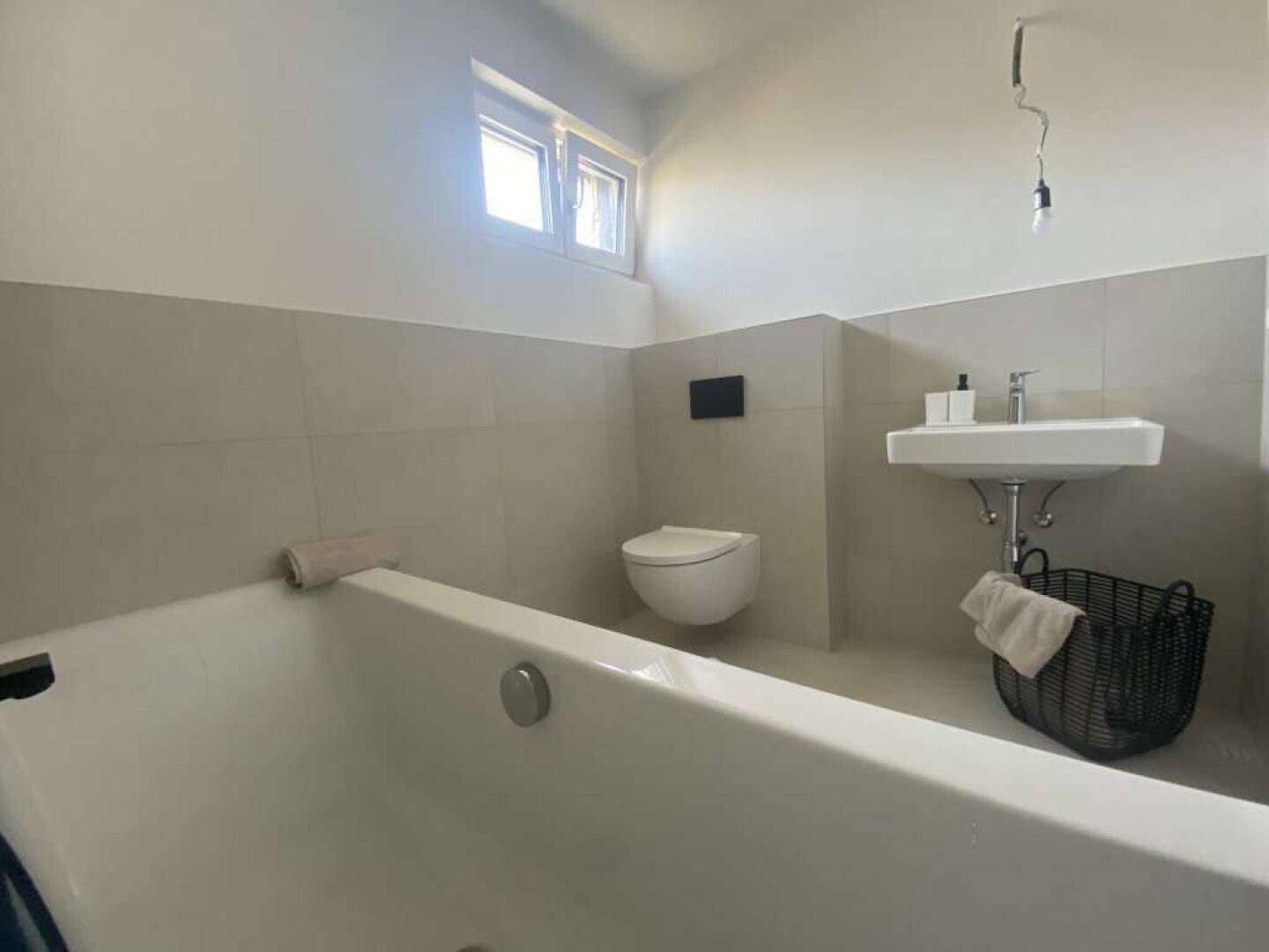 Helles Badezimmer mit Badewanne, Wand-WC, Waschbecken und einem Fenster für Tageslicht.