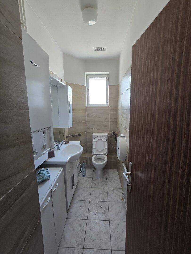 Modernes Badezimmer mit WC, Waschbecken, Spiegelschrank und Fenster, hell gefliest.