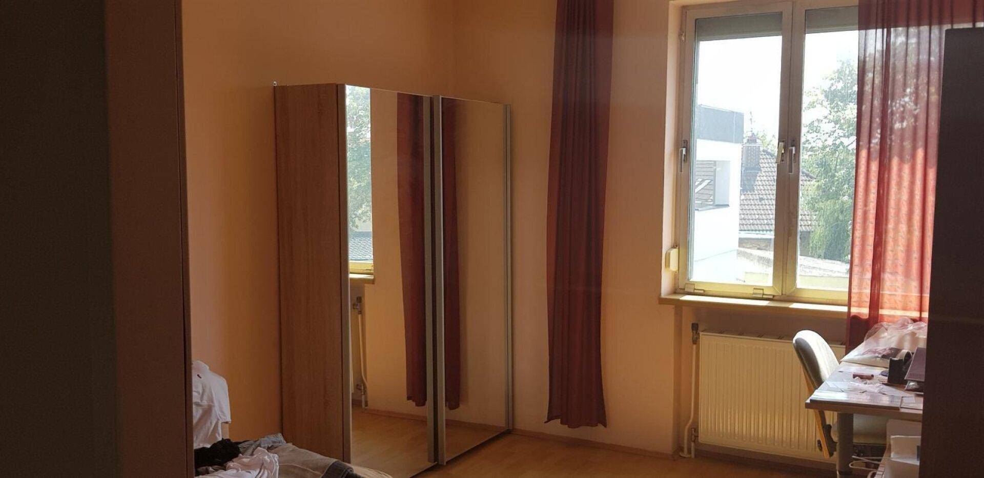 Schlafzimmer mit Spiegelschrank, Schreibtisch am Fenster und roten Vorhängen.