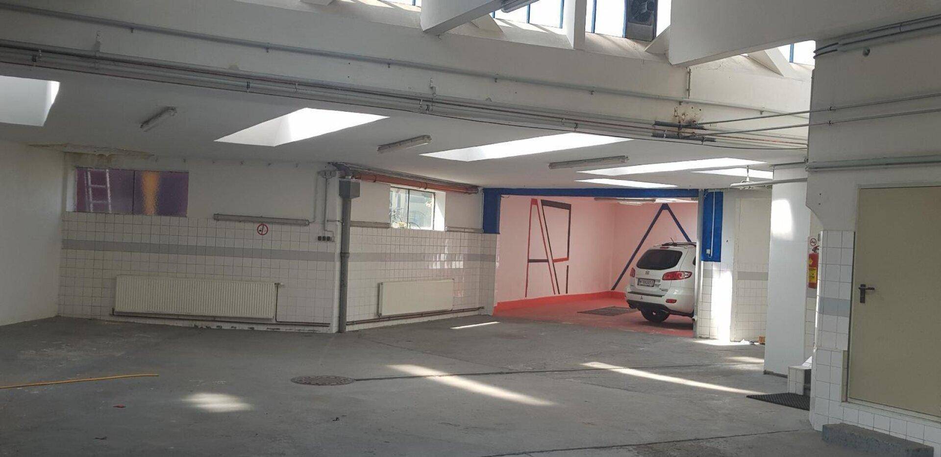 Großer, heller Industrieraum mit Betonboden, Oberlichtern und einem geparkten Auto im Hintergrund.