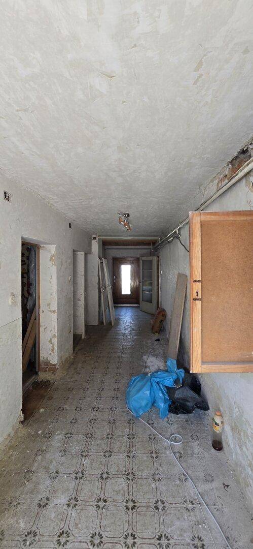 Langer Flur in einem renovierungsbedürftigen Zustand mit alten Fliesen und offener Tür am Ende.