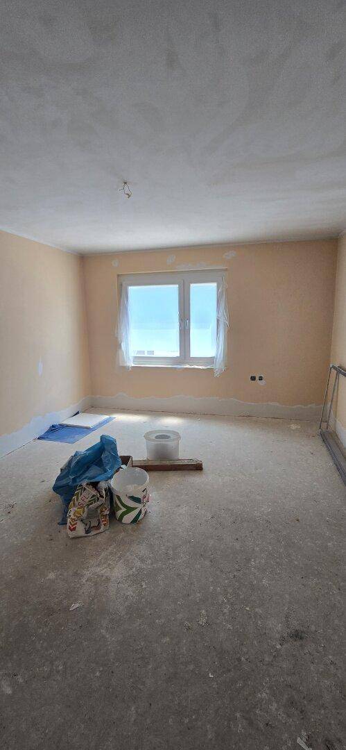 Renovierungsbedürftiges Zimmer mit hellbraunen Wänden, Betonboden und einem Fenster.