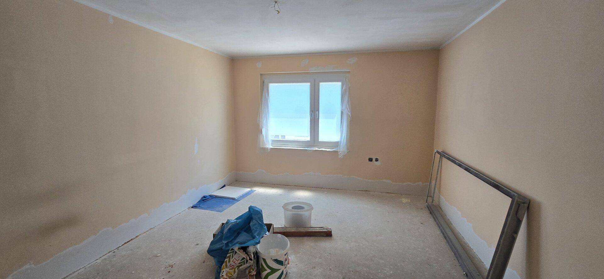 Renovierungsbedürftiges Zimmer mit hellbraunen Wänden, Betonboden und einem Fenster.