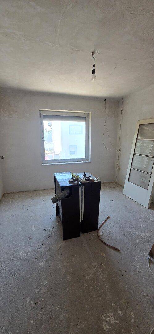 Leerer Raum in Renovierung mit Betonboden, einem Fenster und einer losen Tür an der Wand.