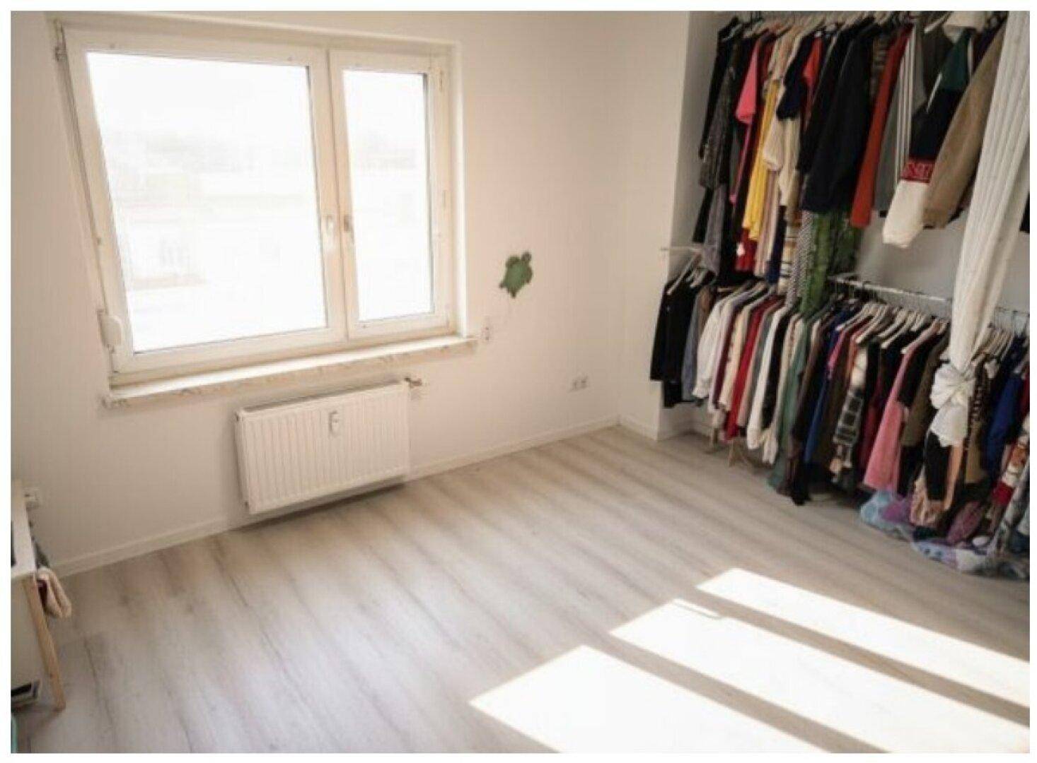 Schlafzimmer mit Kleiderstangen, hellem Laminatboden und großem Fenster für Tageslicht.