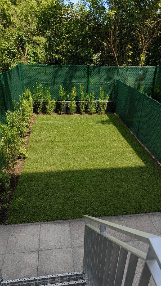 Kleiner, eingezäunter Gartenbereich mit Kunstrasen und Metalltreppe, umgeben von Bäumen.