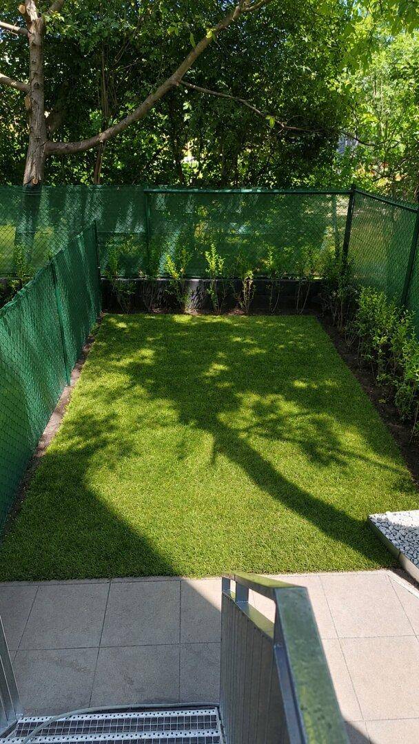 Kleiner, eingezäunter Gartenbereich mit Kunstrasen und Metalltreppe, umgeben von Bäumen.
