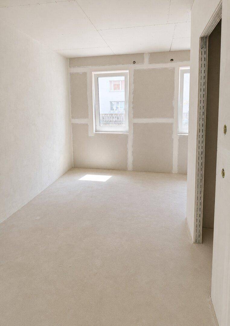 Unfertiger Raum mit Trockenbauwänden, hellem Estrichboden und zwei Fenstern, bereit für den weiteren Ausbau.