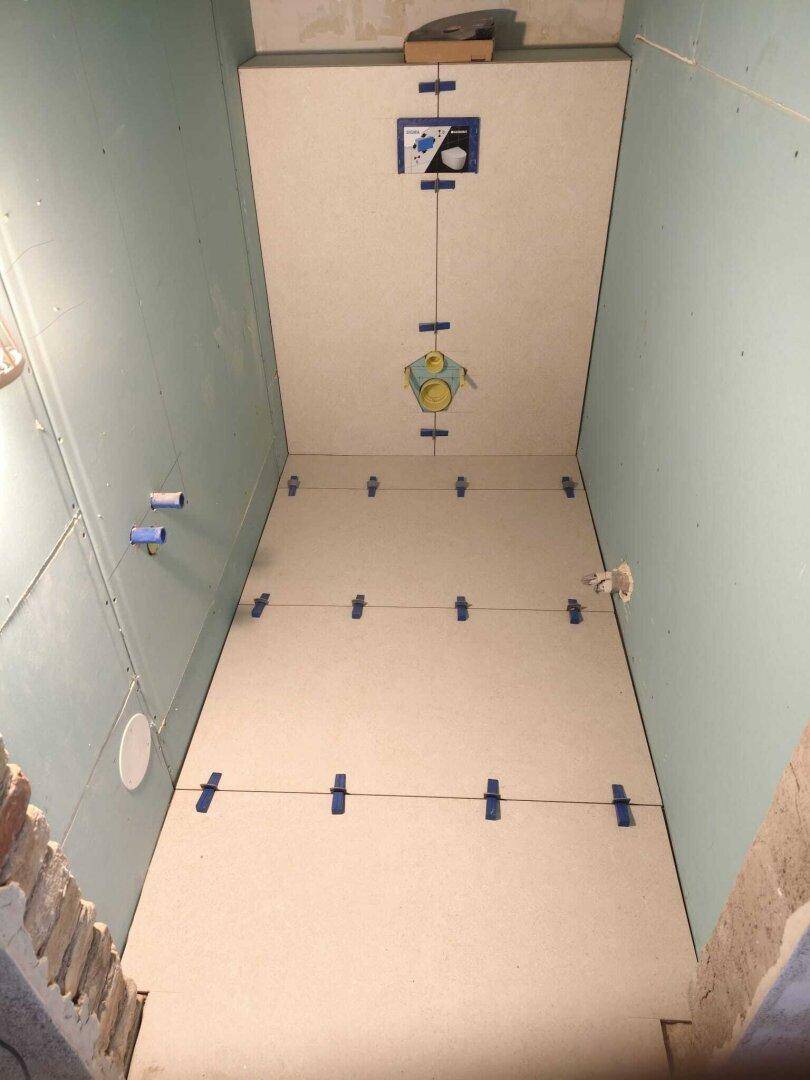 Badezimmer im Bau mit Fliesenarbeiten am Boden, blauen Trockenbauwänden und Installationen für die Toilette.