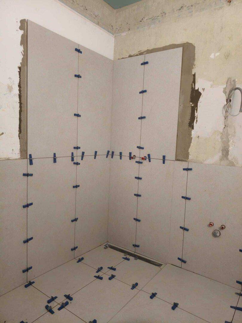 Badezimmer im Bau mit Fliesenarbeiten an Wänden und Boden, inklusive Fugenkreuzen und Duschablauf.