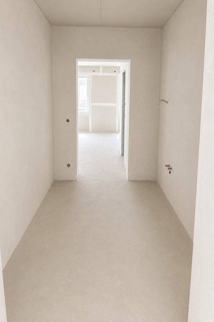 Unfertiger Flur oder Eingangsbereich mit hellem Boden und weißen Wänden, der zu einem weiteren Raum führt.