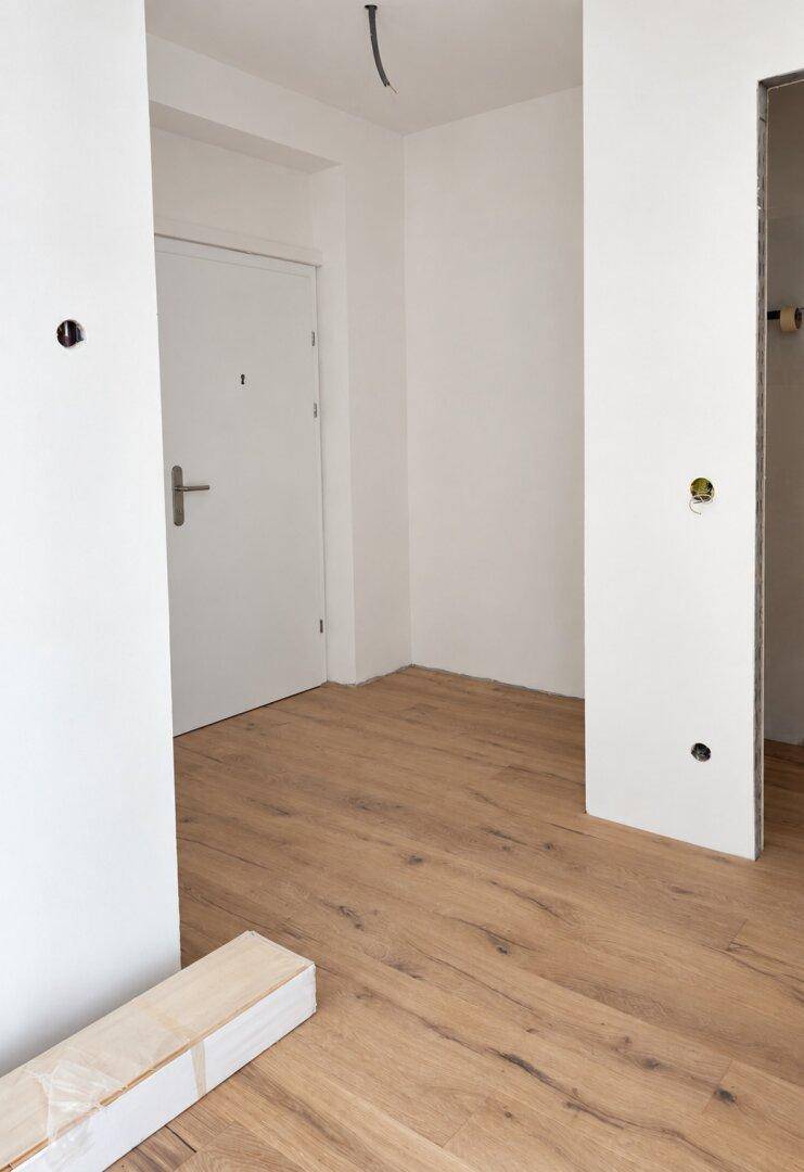 Eingangsbereich im Rohbau mit weißer Eingangstür und neu verlegtem hellem Holzplankenboden.