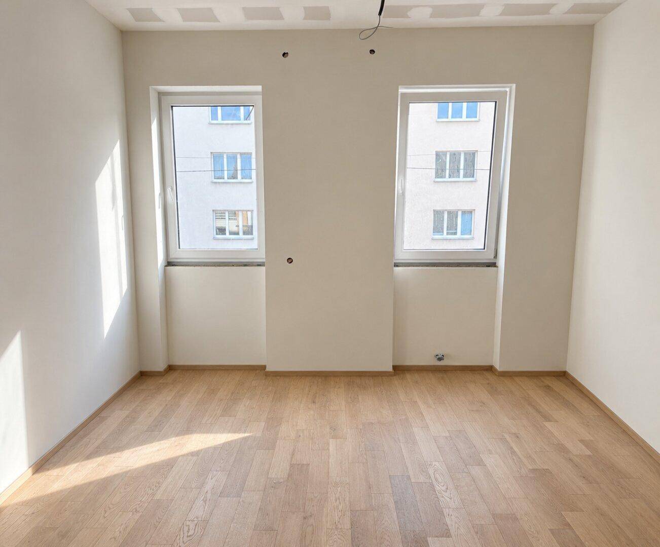 Heller Raum im Rohbau mit zwei großen Fenstern, die viel Tageslicht hereinlassen und auf ein Gebäude blicken.