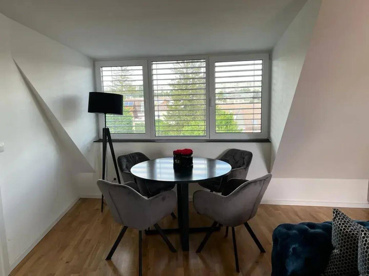 Heller Essbereich mit modernem runden Tisch, grauen Stühlen und Fenster mit Jalousien.