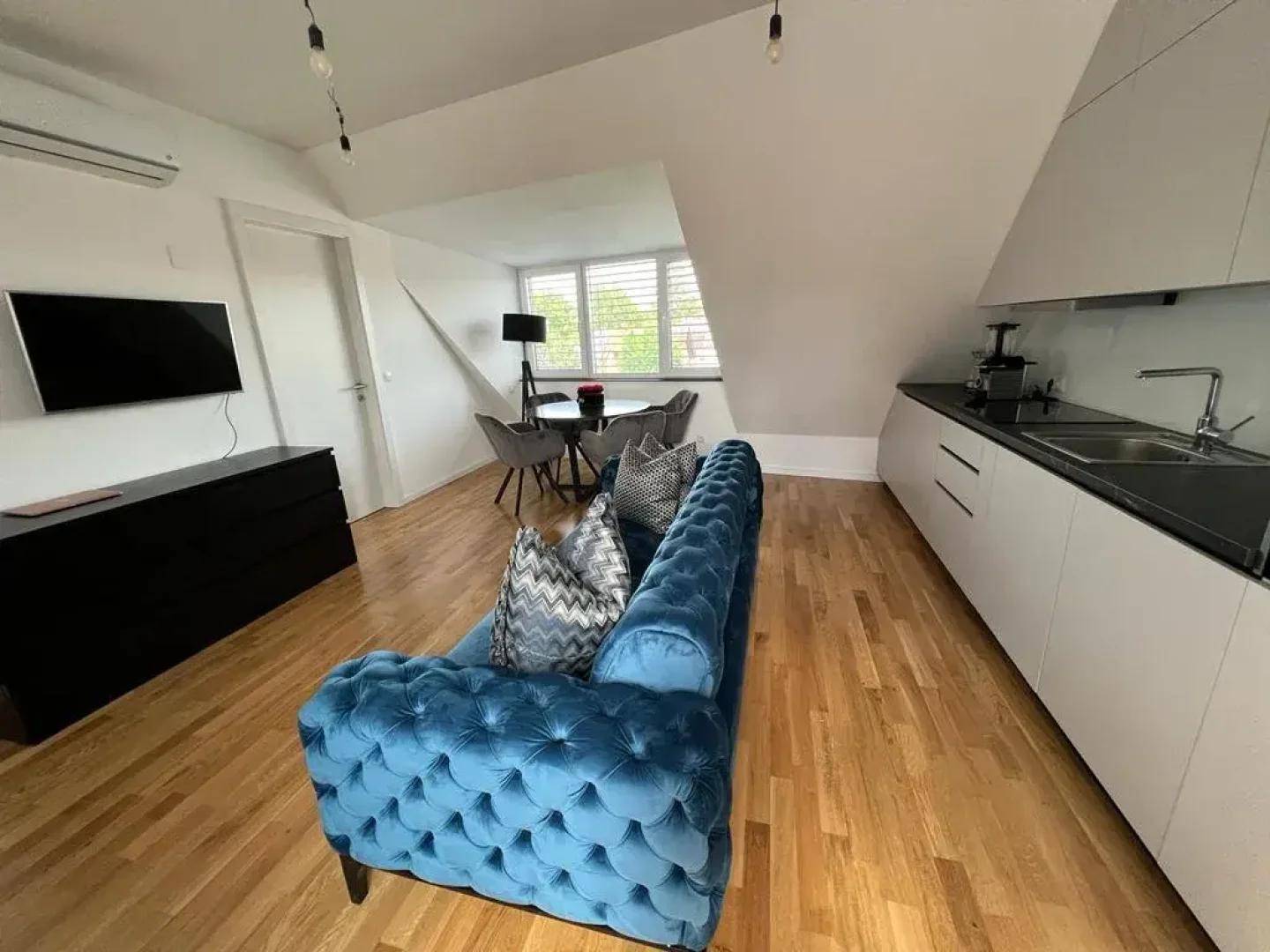 Heller Wohnbereich mit offener Küche, blauer Couch und Essbereich unter Dachschräge.