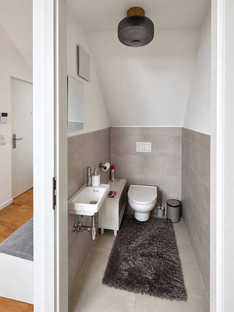 Kleines Gäste-WC mit wandhängendem WC, kleinem Waschbecken und grauen Fliesen.