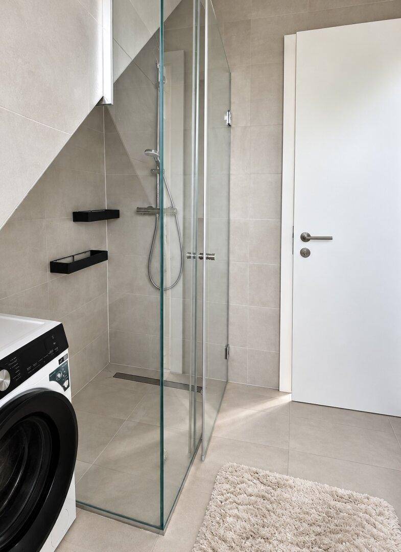 Modernes Badezimmer mit begehbarer Dusche, grauen Fliesen und Waschmaschine.