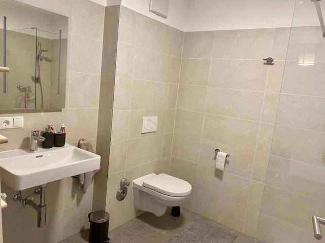 Helles Badezimmer mit Wand-WC, Waschbecken und großformatigen beigefarbenen Fliesen.