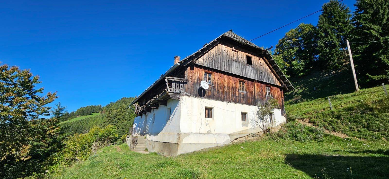 Rustikales Holzhaus mit weiß getünchtem Sockel und traditionellem Dach in grüner Hügellandschaft.