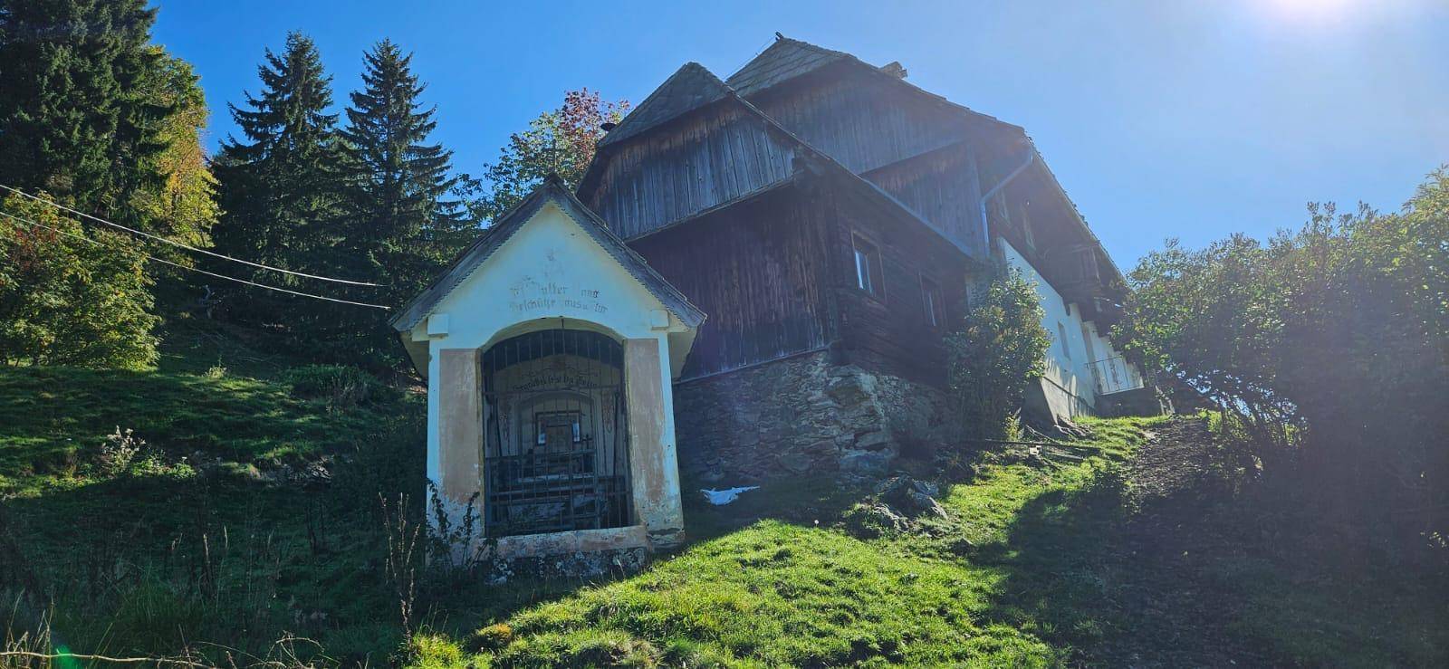 Historisches Bauernhaus mit kleiner Kapelle auf einem sonnigen Hügel in ländlicher Umgebung.