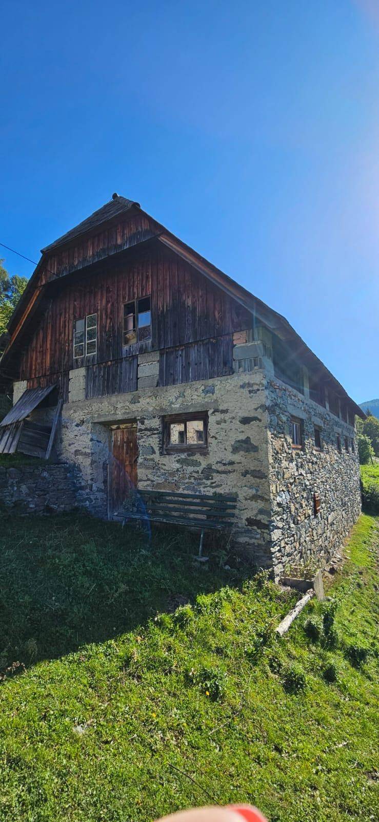 Altes Bauernhaus mit Steinsockel und Holzfassade, umgeben von grüner Natur.