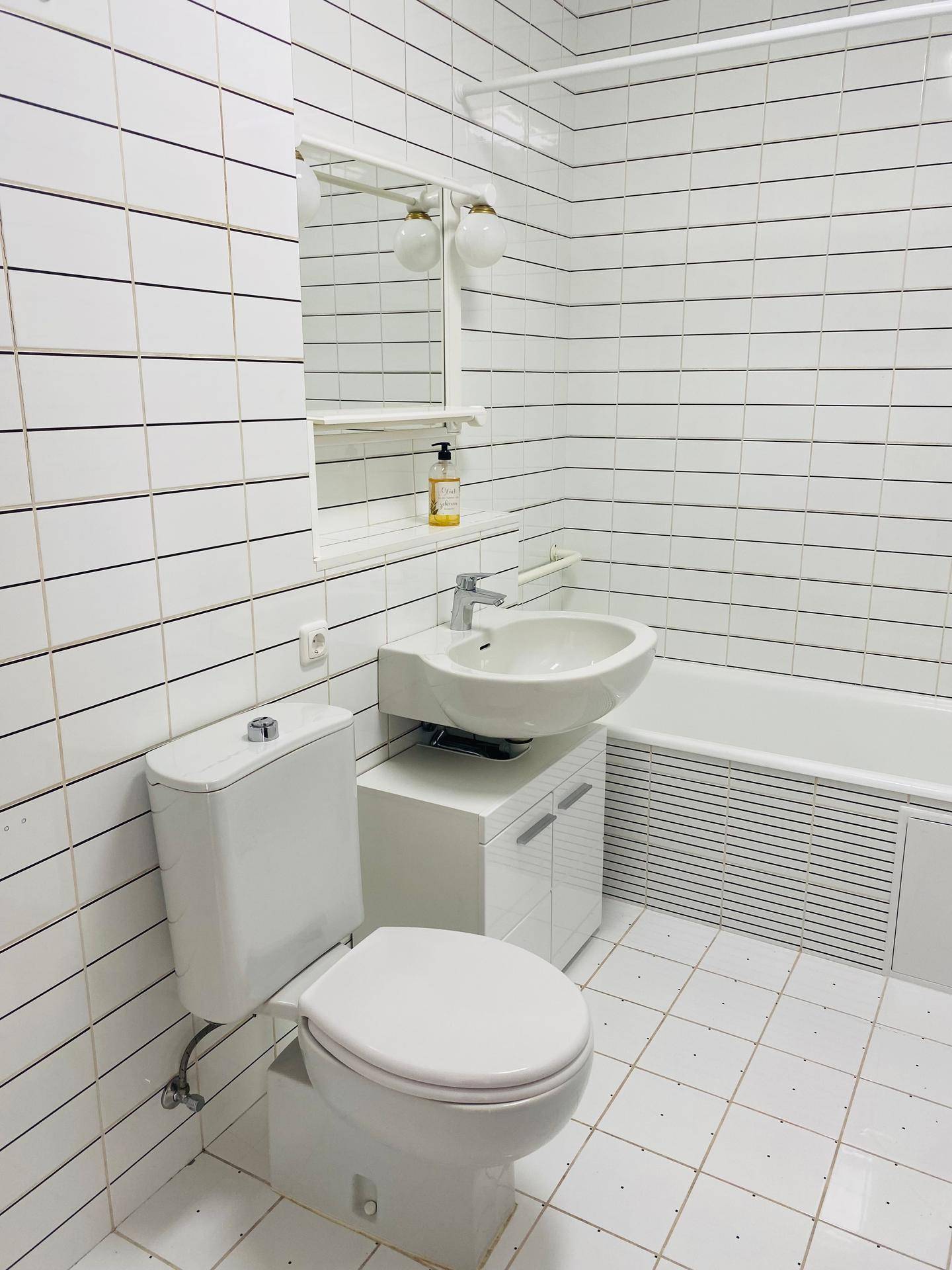 Funktionales Badezimmer mit weißen Fliesen, WC, Waschbecken mit Unterschrank und Badewanne.