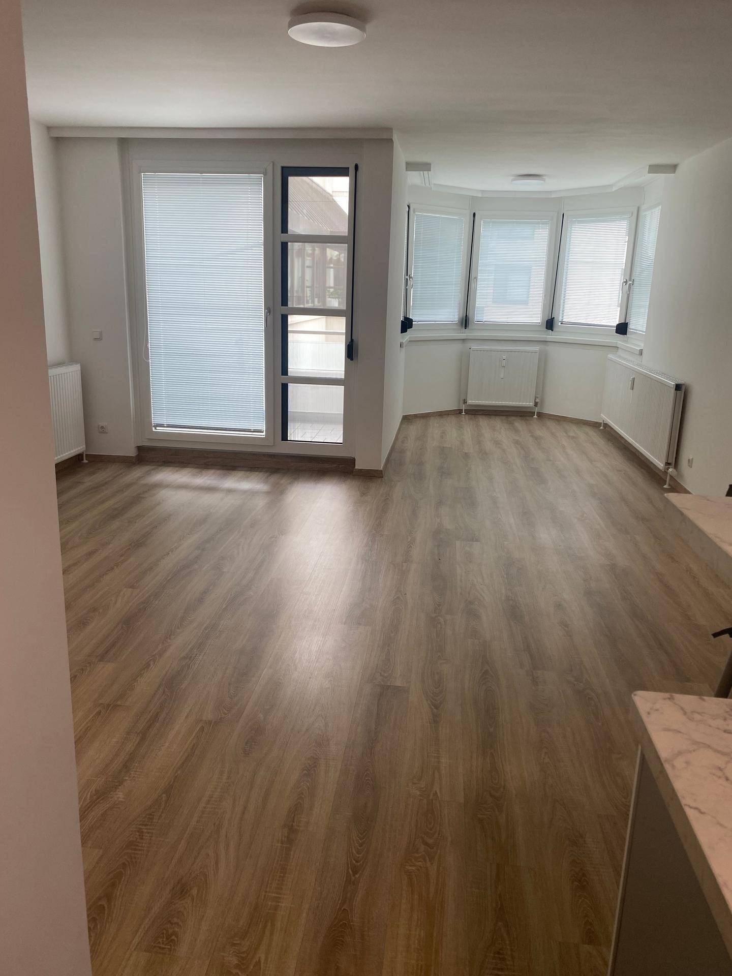 Geräumiges Wohnzimmer mit Laminatboden, großen Fenstern mit Jalousien und Balkonzugang.