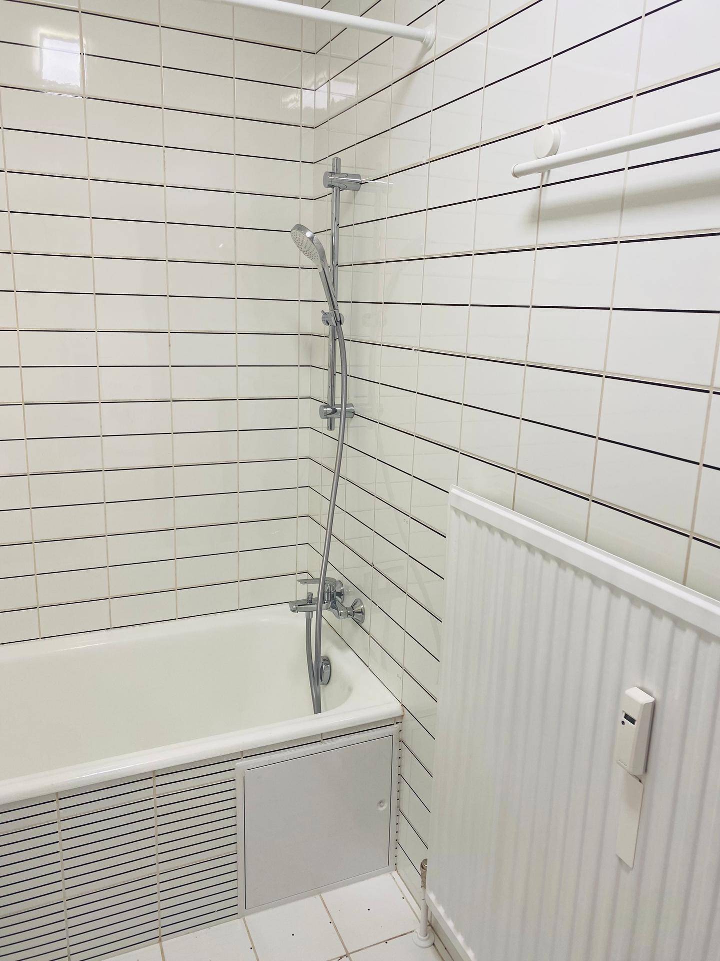 Badewanne mit Duscharmatur und Heizkörper in einem hell gefliesten Badezimmer.
