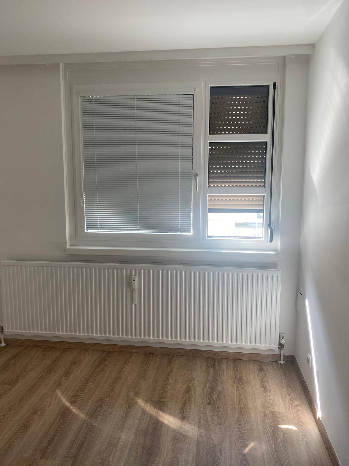 Heller Raum mit Laminatboden, Fenster mit Jalousien und einem Heizkörper unter dem Fenster.