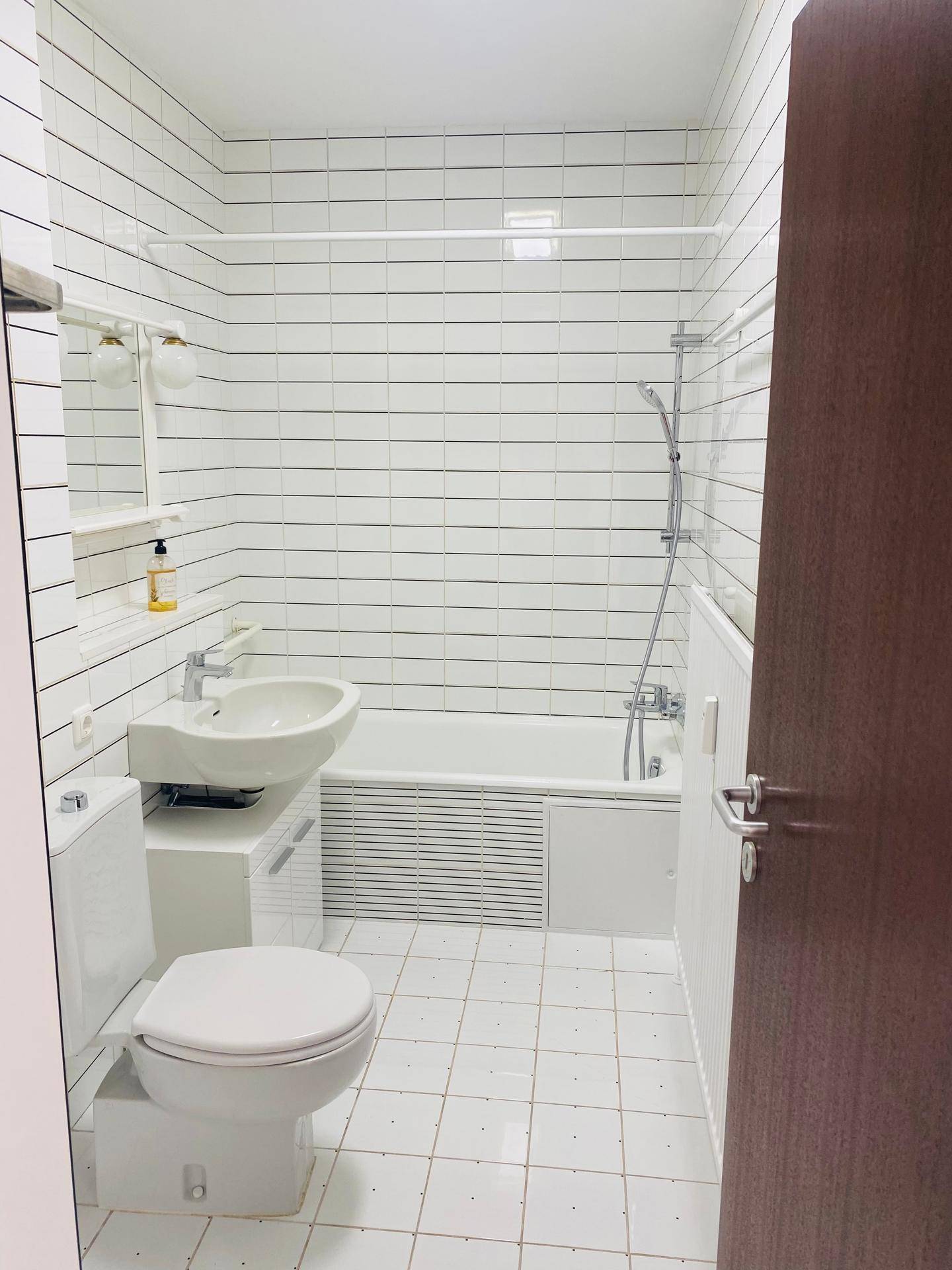 Helles Badezimmer mit weißen Fliesen, Toilette, Waschbecken und einer Badewanne mit Duschfunktion.