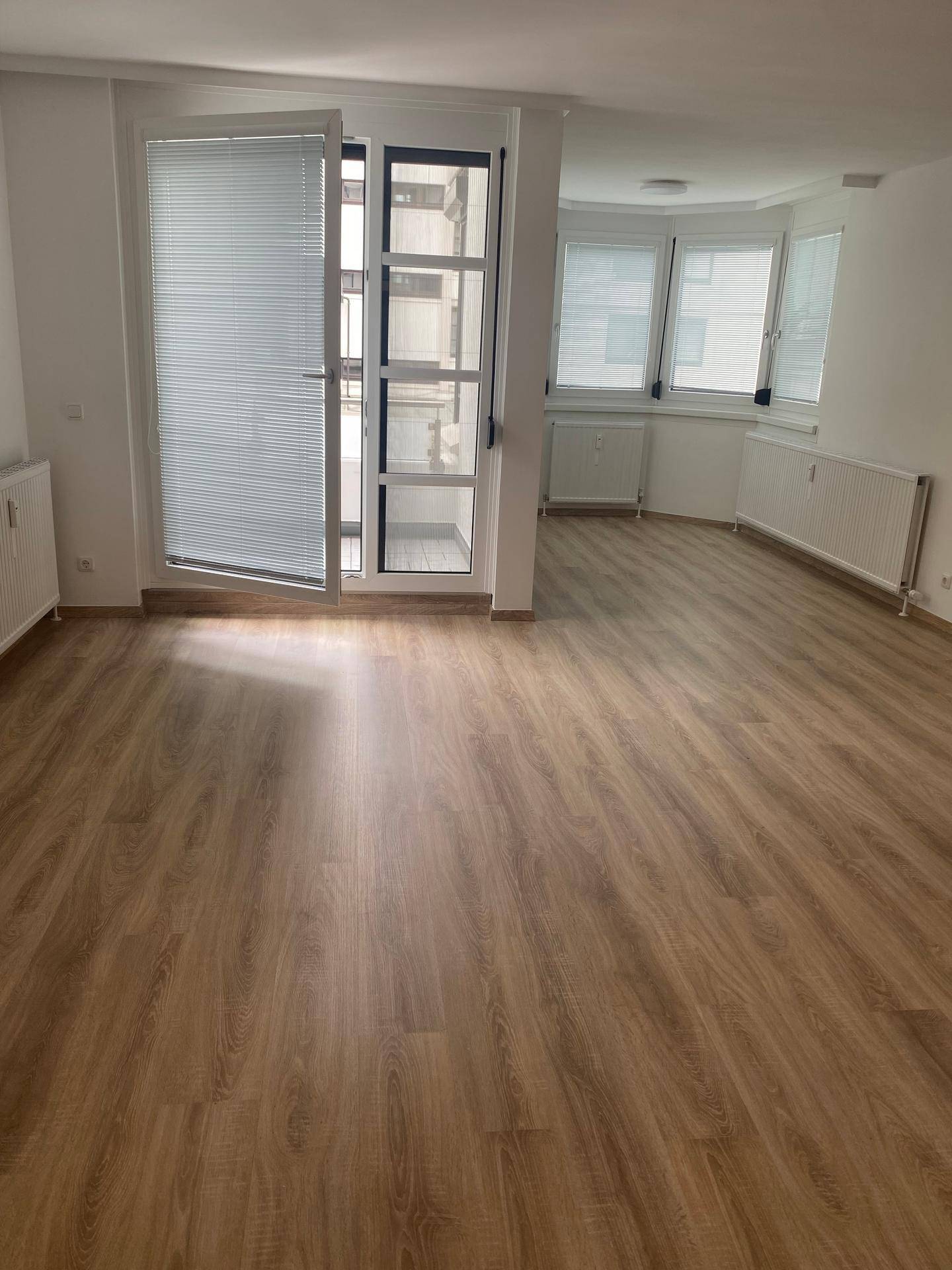 Helles Wohnzimmer mit Laminatboden, großen Fenstern mit Jalousien und Zugang zum Balkon.