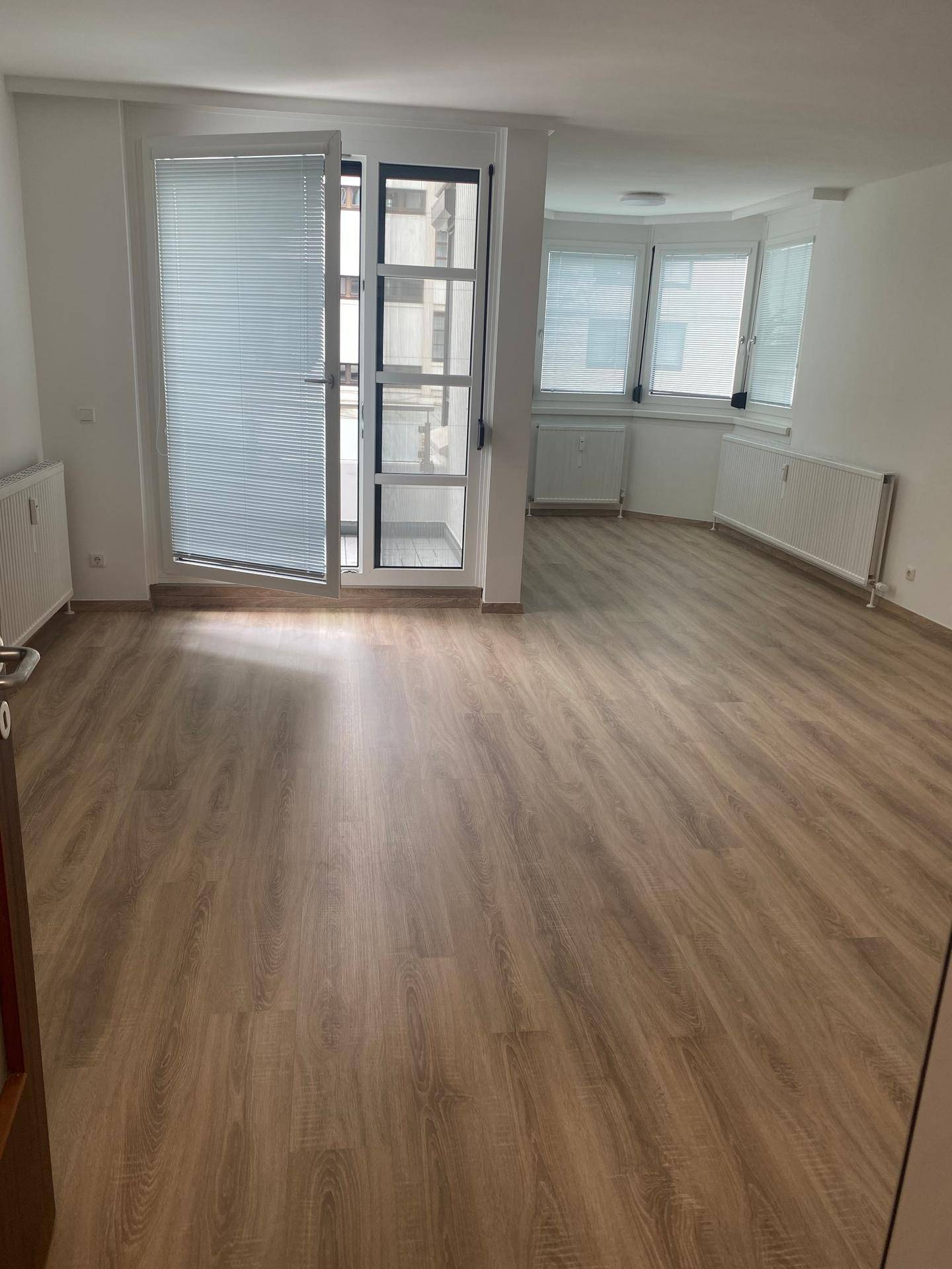Heller, leerer Wohnraum mit modernem Laminatboden, großen Fenstern und direktem Zugang zum Balkon.