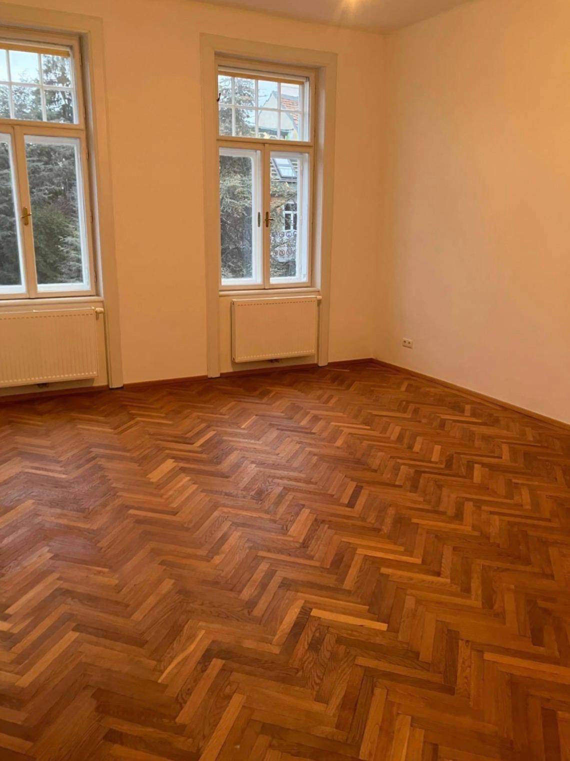 Geräumiges Zimmer mit klassischem Fischgrätparkett und zwei hohen Fenstern für viel Tageslicht.