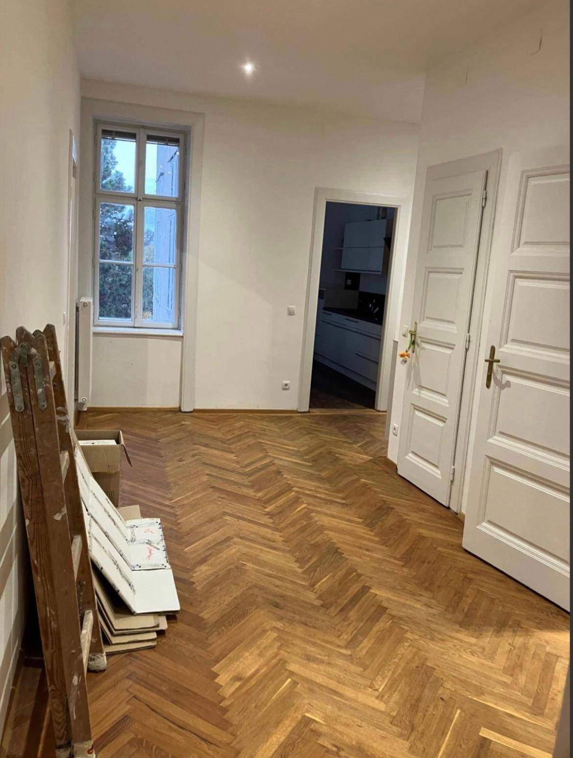 Leeres Zimmer mit Fischgrätparkett, einem Fenster und Blick in die angrenzende Küche.