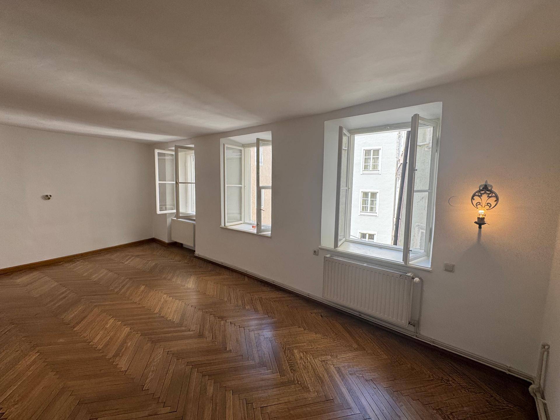 Helles Zimmer mit Fischgrätparkett, hohen Fenstern und Blick auf die umliegenden Gebäude.