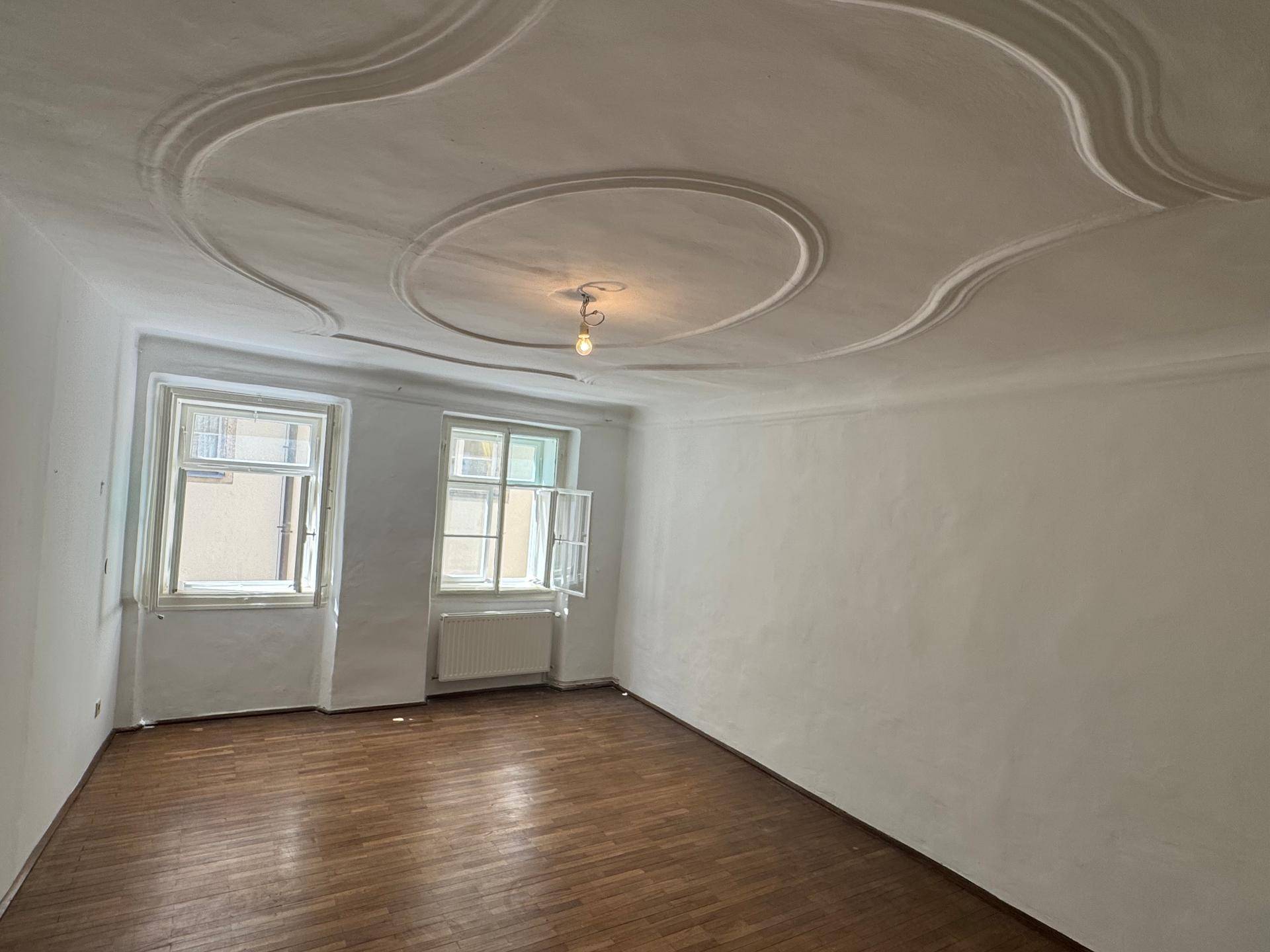 Geräumiges Zimmer mit Holzboden, dekorativer Stuckdecke und zwei großen Fenstern.