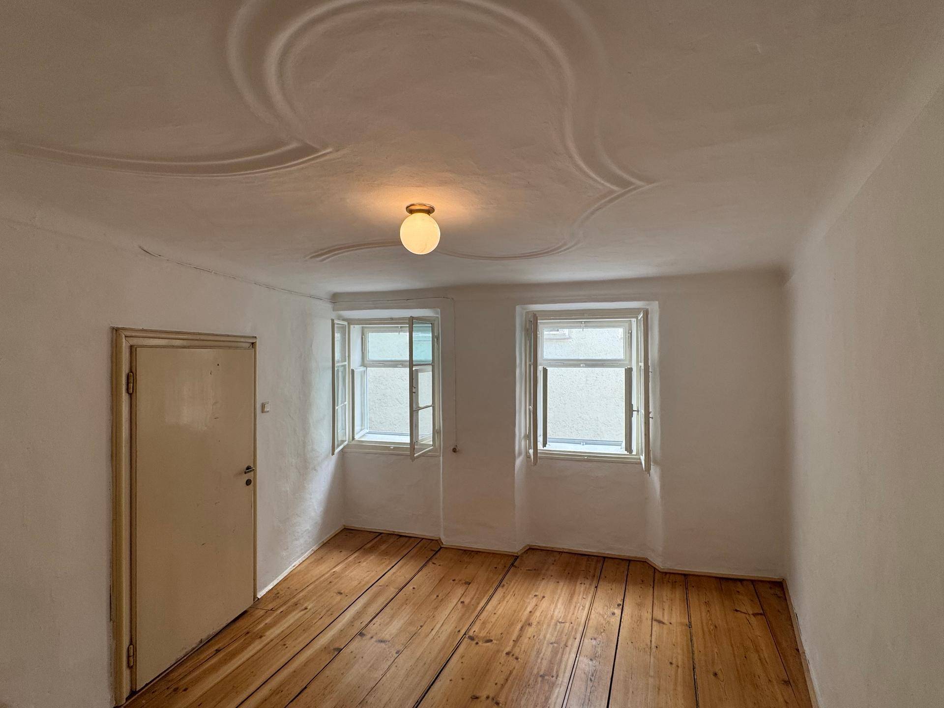 Helles Zimmer mit breiten Holzplanken, dekorativer Stuckdecke und zwei Fenstern.