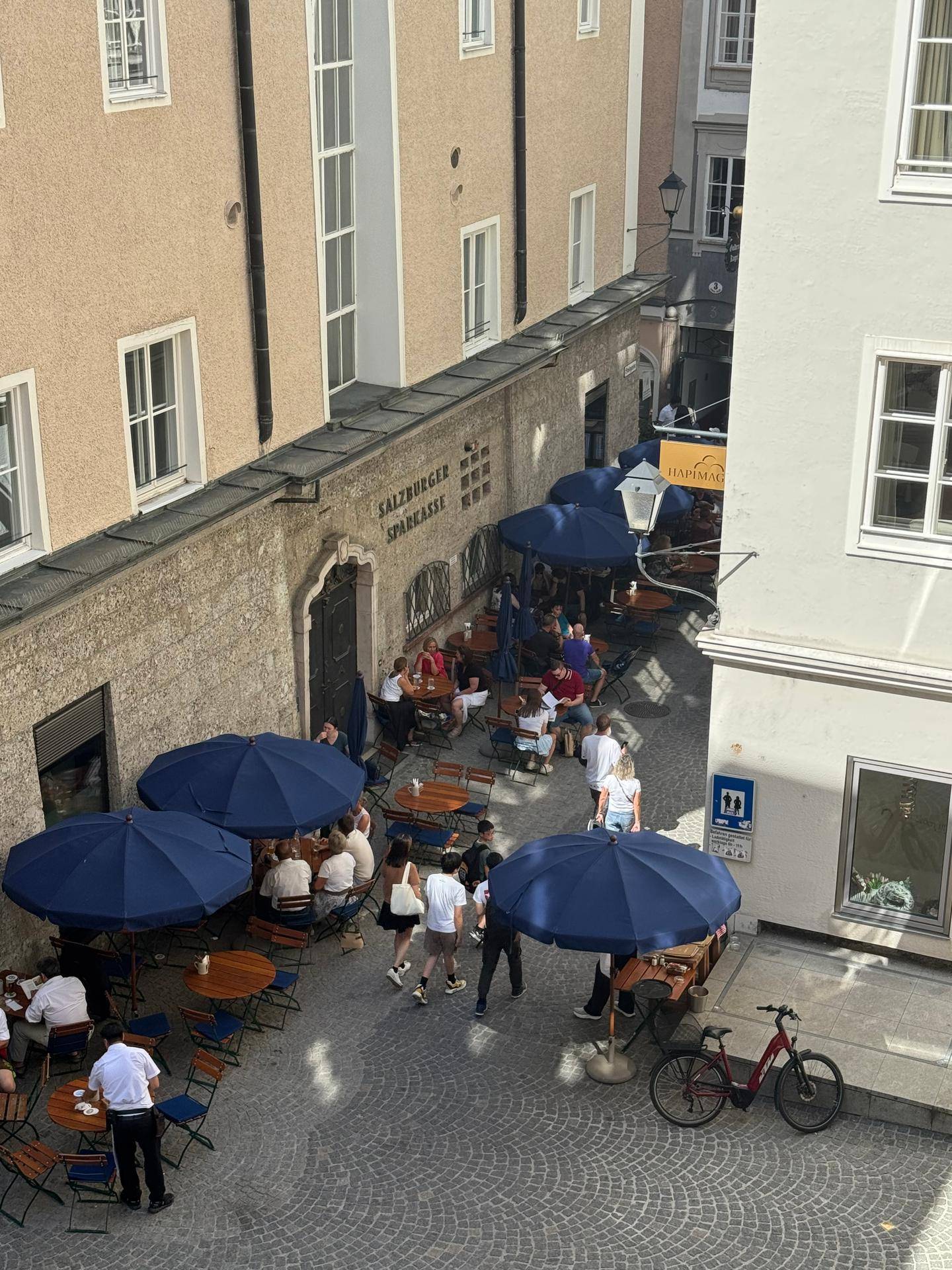 Blick von oben auf eine belebte Gasse mit Cafés und Sonnenschirmen in der Altstadt.