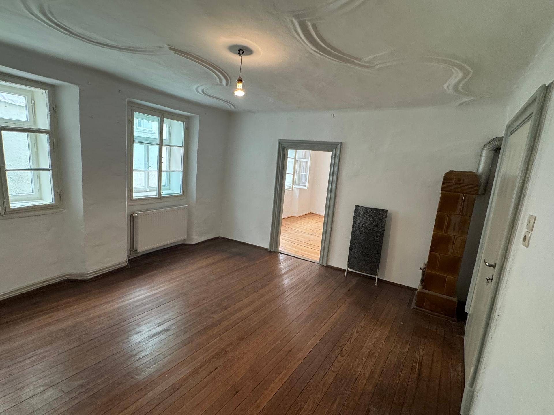 Geräumiges Zimmer mit Holzboden, Stuckdecke und einem traditionellen Kachelofen.