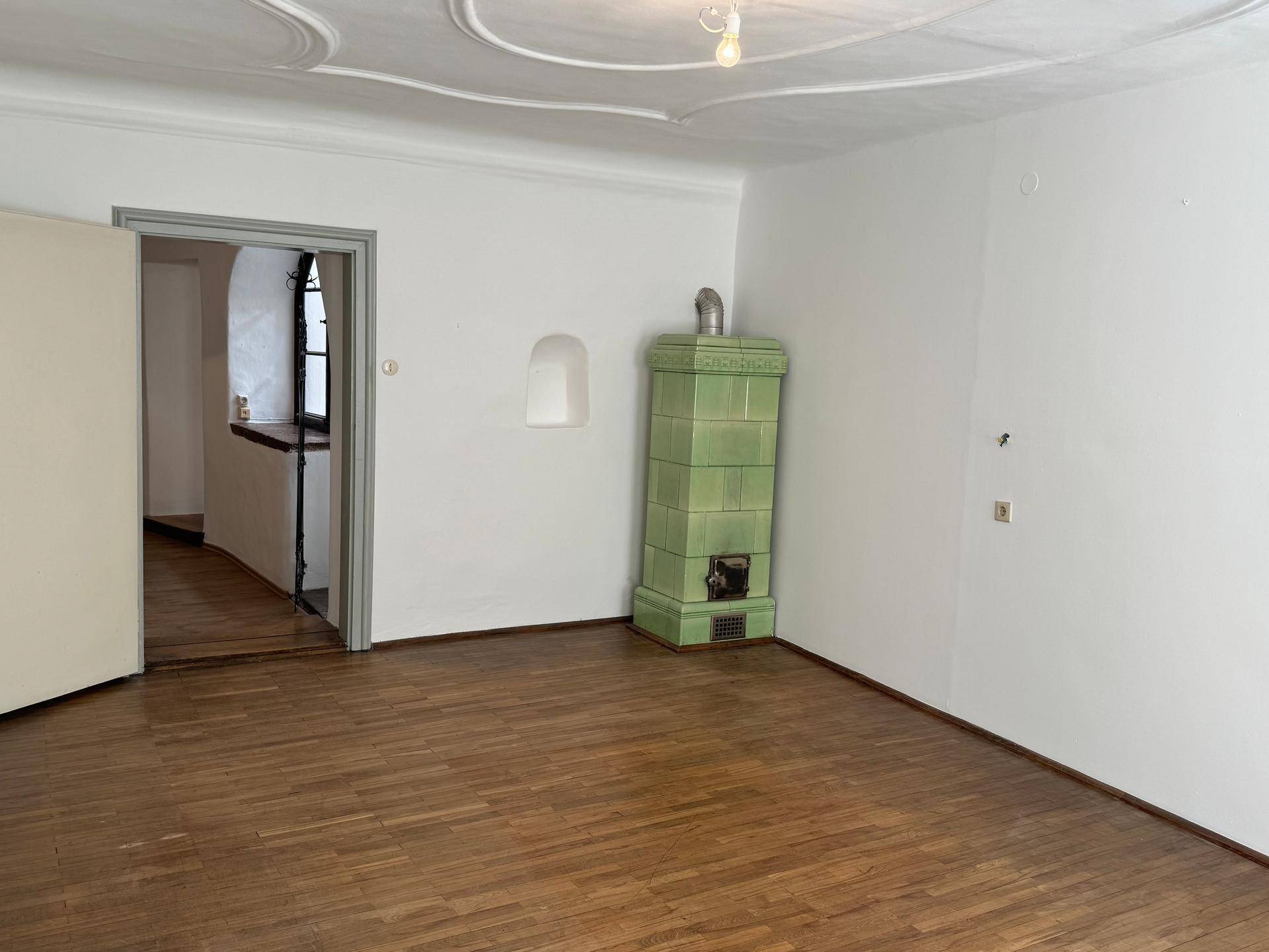 Heller Raum mit Holzboden und einem traditionellen grünen Kachelofen als Blickfang.