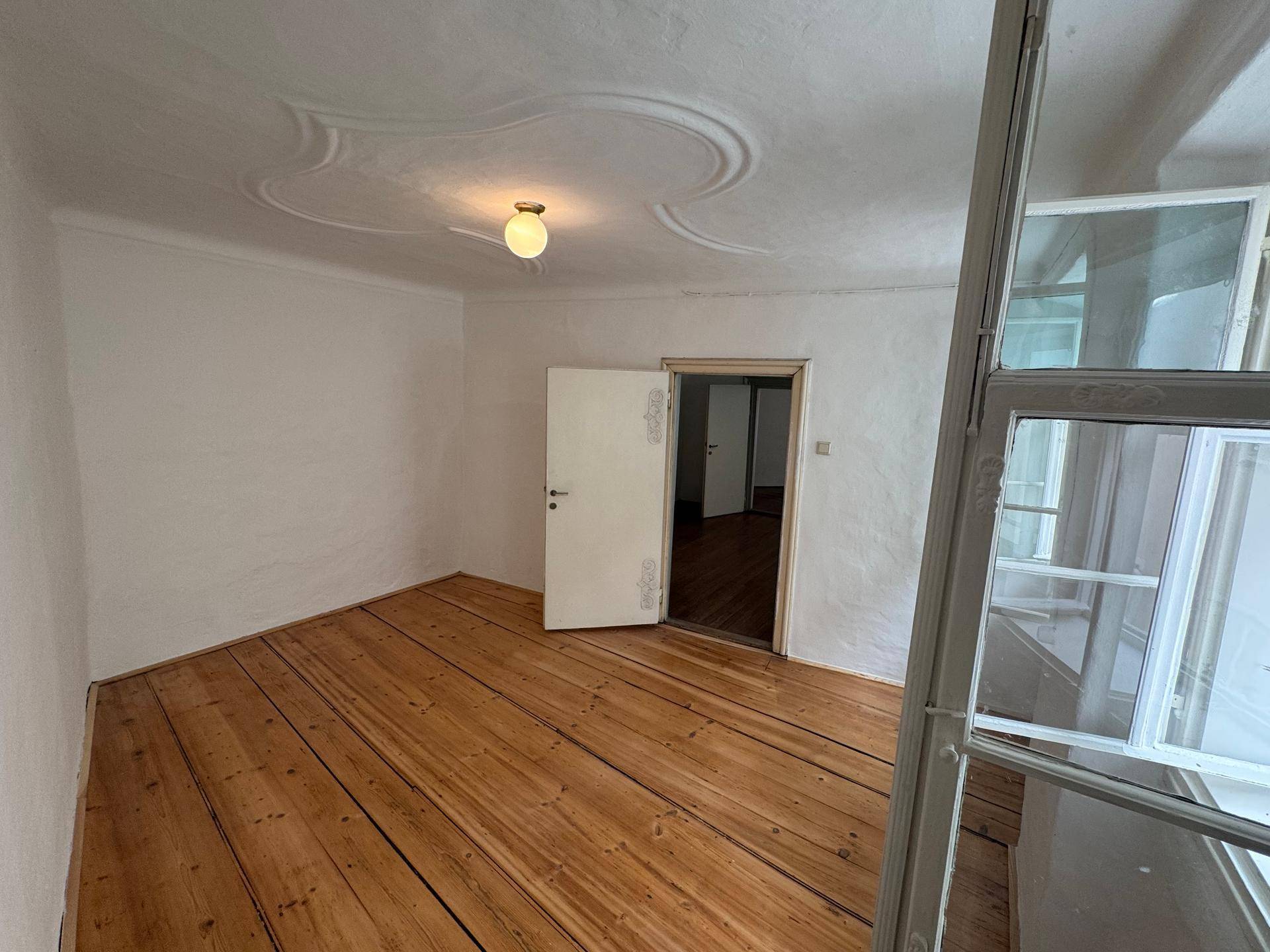 Heller Raum mit breiten Holzplanken, Stuckdecke und Blick durch ein geöffnetes Fenster.
