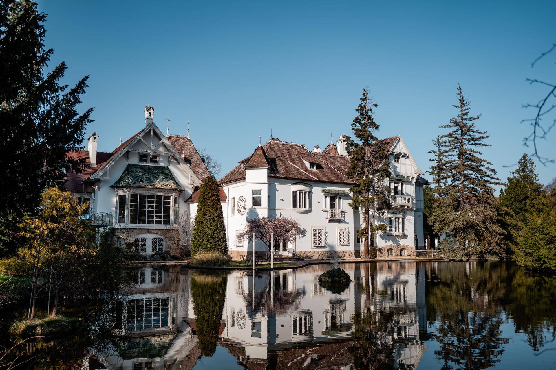 Historisches Gebäude mit weißer Fassade und roten Dächern, gespiegelt in einem ruhigen Teich.