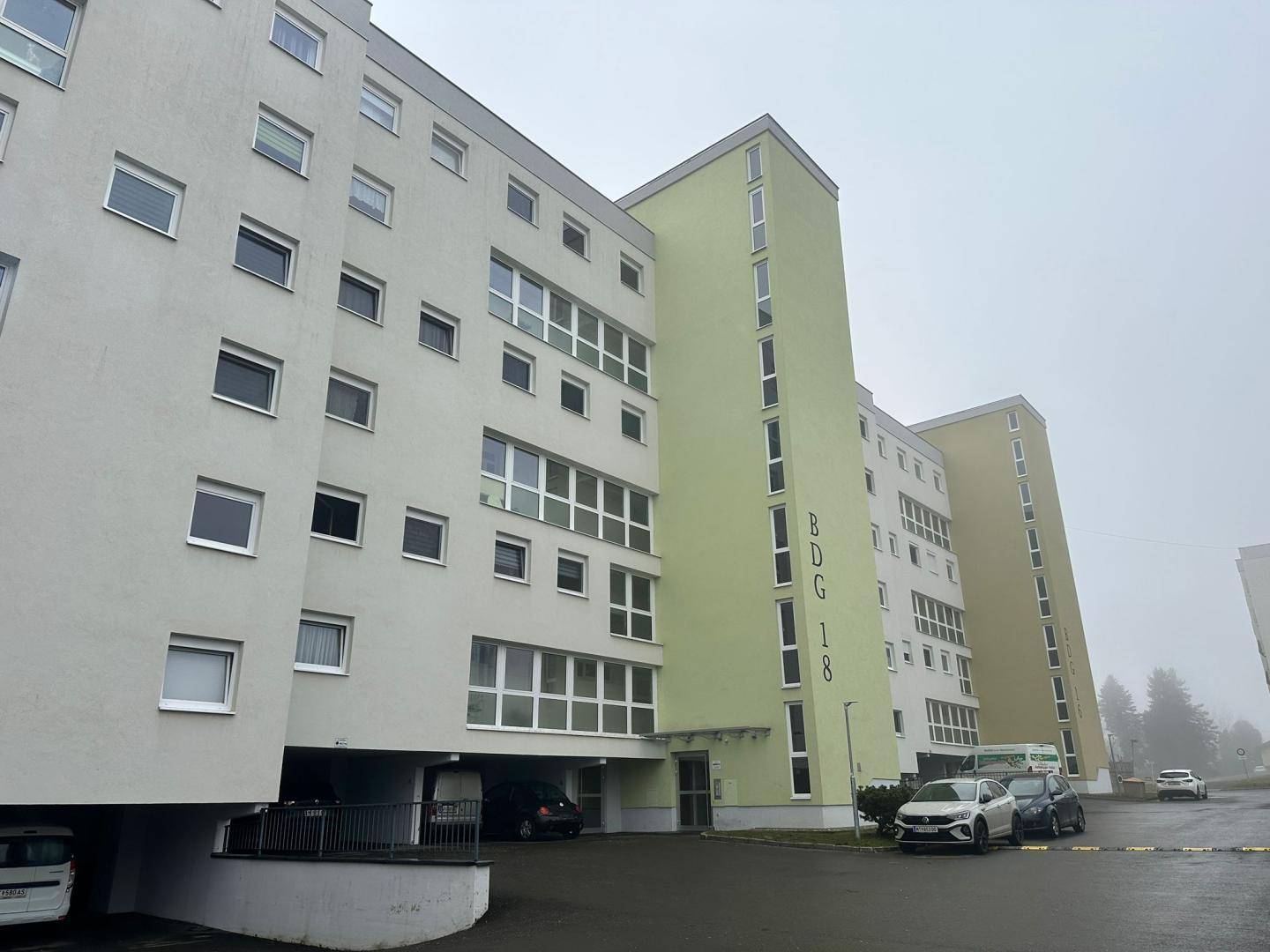 Mehrstöckiges Wohngebäude mit hellgrüner und beiger Fassade sowie Parkplätzen im Vordergrund.